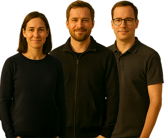 Drei Personen stehen nebeneinander und lächeln in die Kamera. Sie tragen dunkle, legere Kleidung und stehen vor einem schlichten, bräunlichen Hintergrund.