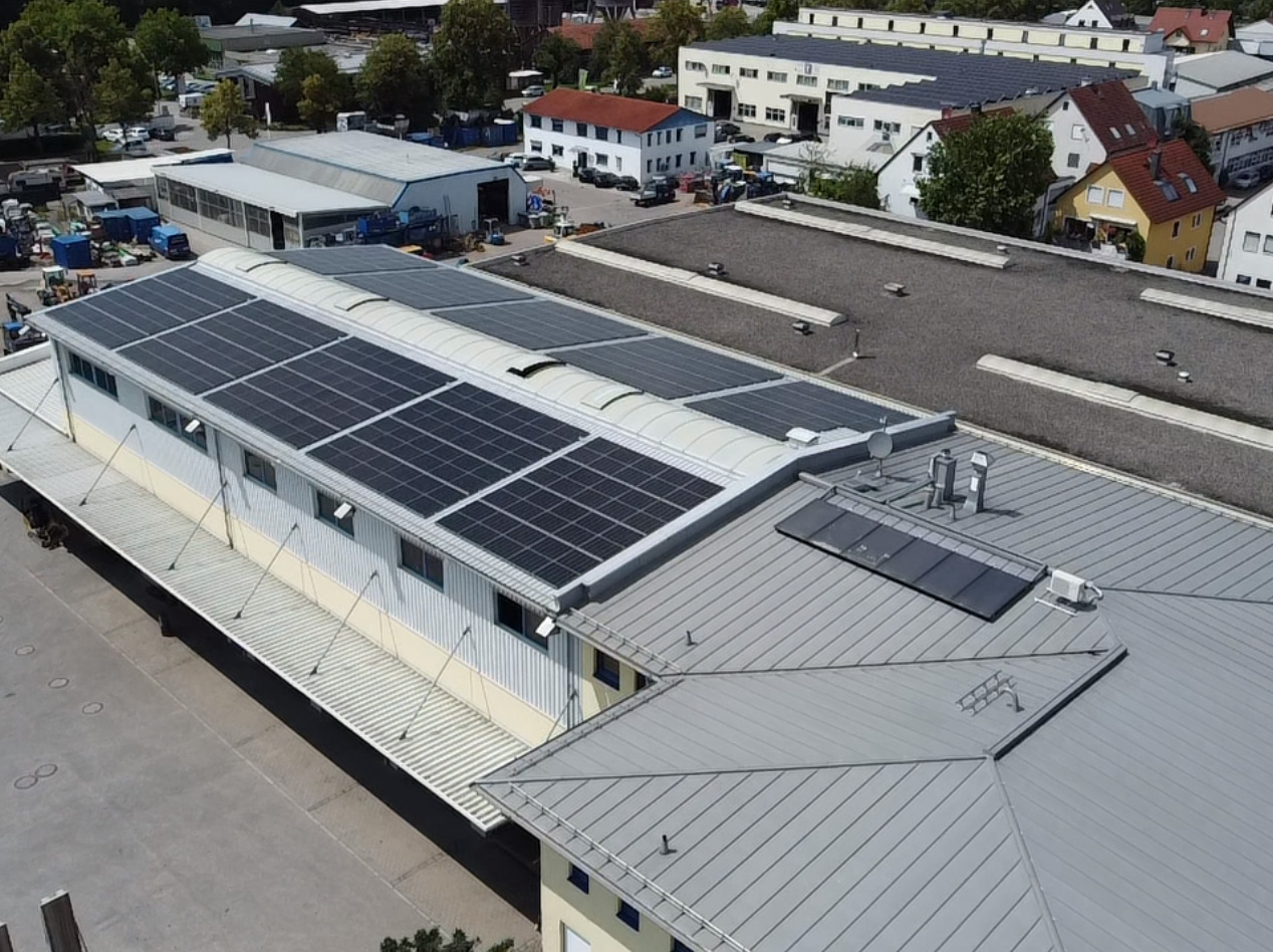 dachau Ein großes Industriegebäude mit zahlreichen auf dem Dach installierten Solarzellen, umgeben von anderen Gebäuden, geparkten Fahrzeugen und einigen Bäumen im Hintergrund.