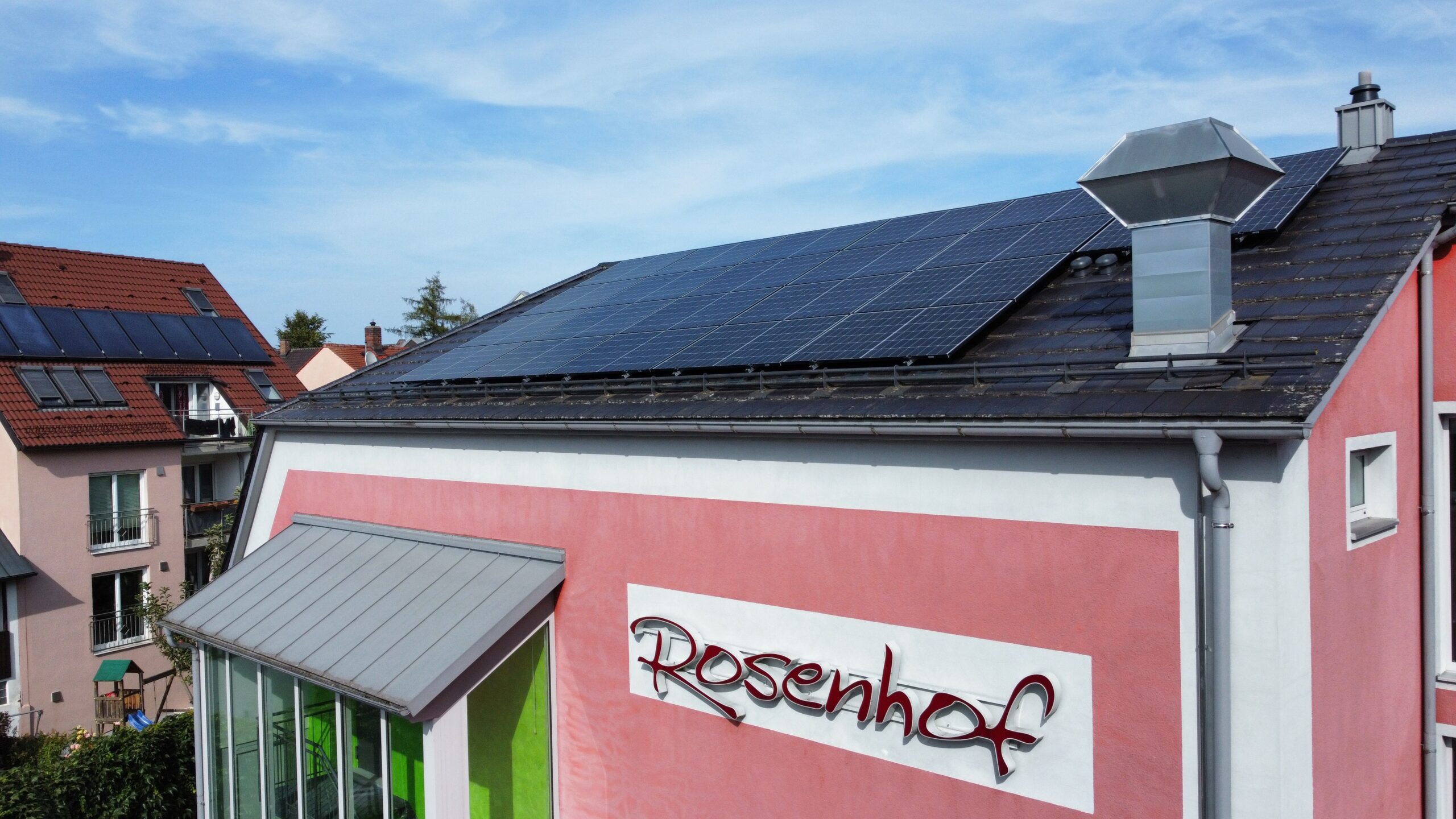 DCIM100MEDIADJI_0725.JPG Ein rosafarbenes Gebäude mit dem Zeichen Rosenhof hat auf seinem Schrägdach Sonnenkollektoren installiert. Im Hintergrund sind unter blauem Himmel weitere Häuser mit ähnlichen Sonnenkollektoren zu sehen.