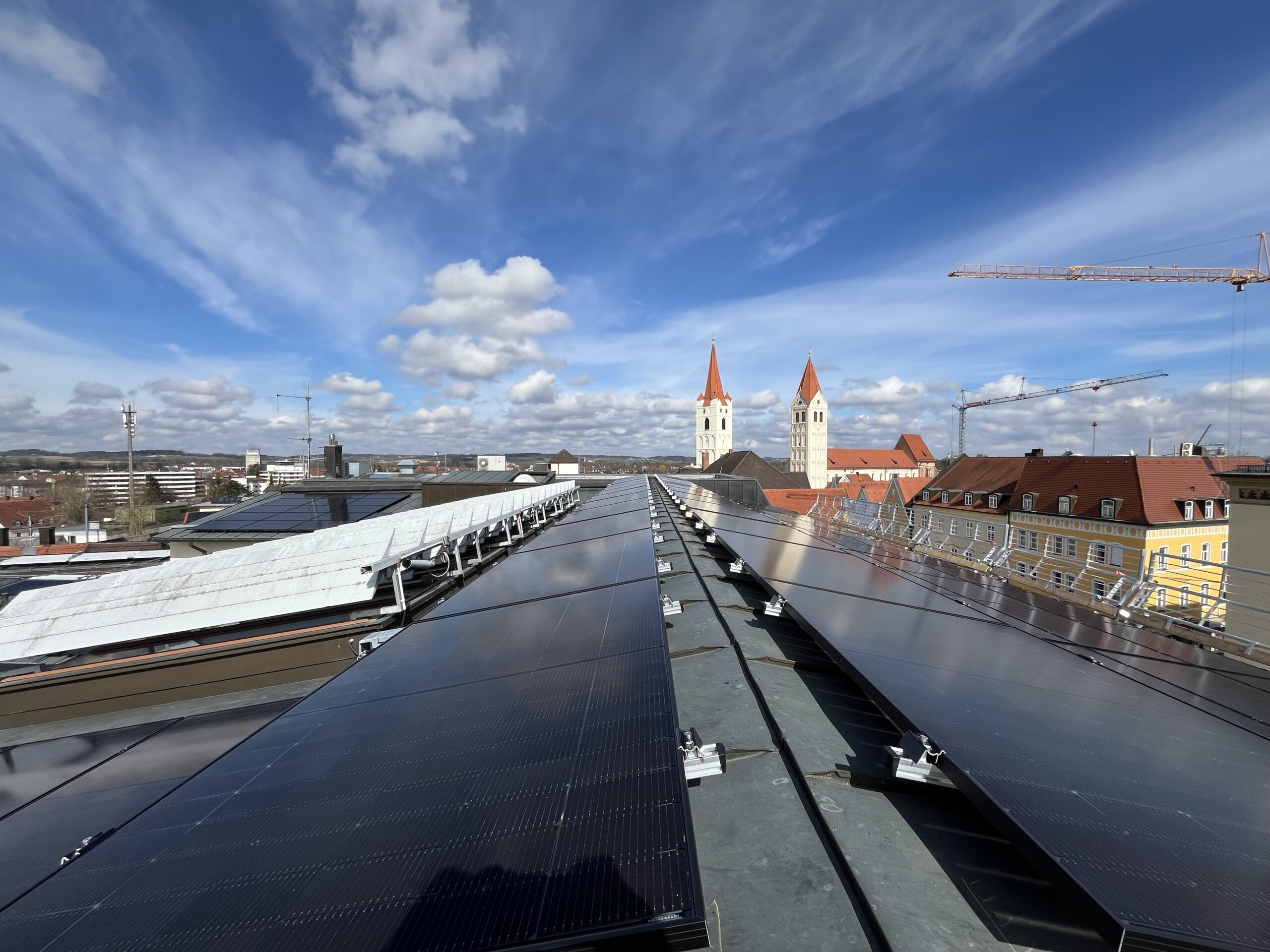 moosburg Sonnenkollektoren auf dem Dach unter einem blauen Himmel mit vereinzelten Wolken; in der Ferne sind zwei weiße Kirchtürme mit roten Dächern zu sehen, umgeben von städtischen Gebäuden und Baukränen.