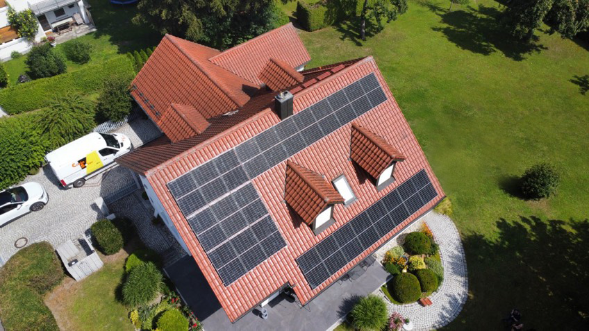niederbayern Luftaufnahme eines Hauses mit rotem Ziegeldach, das mit Solarzellen bedeckt ist, umgeben von grünem Rasen, Gärten, Sträuchern und geparkten Fahrzeugen auf einer Einfahrt.