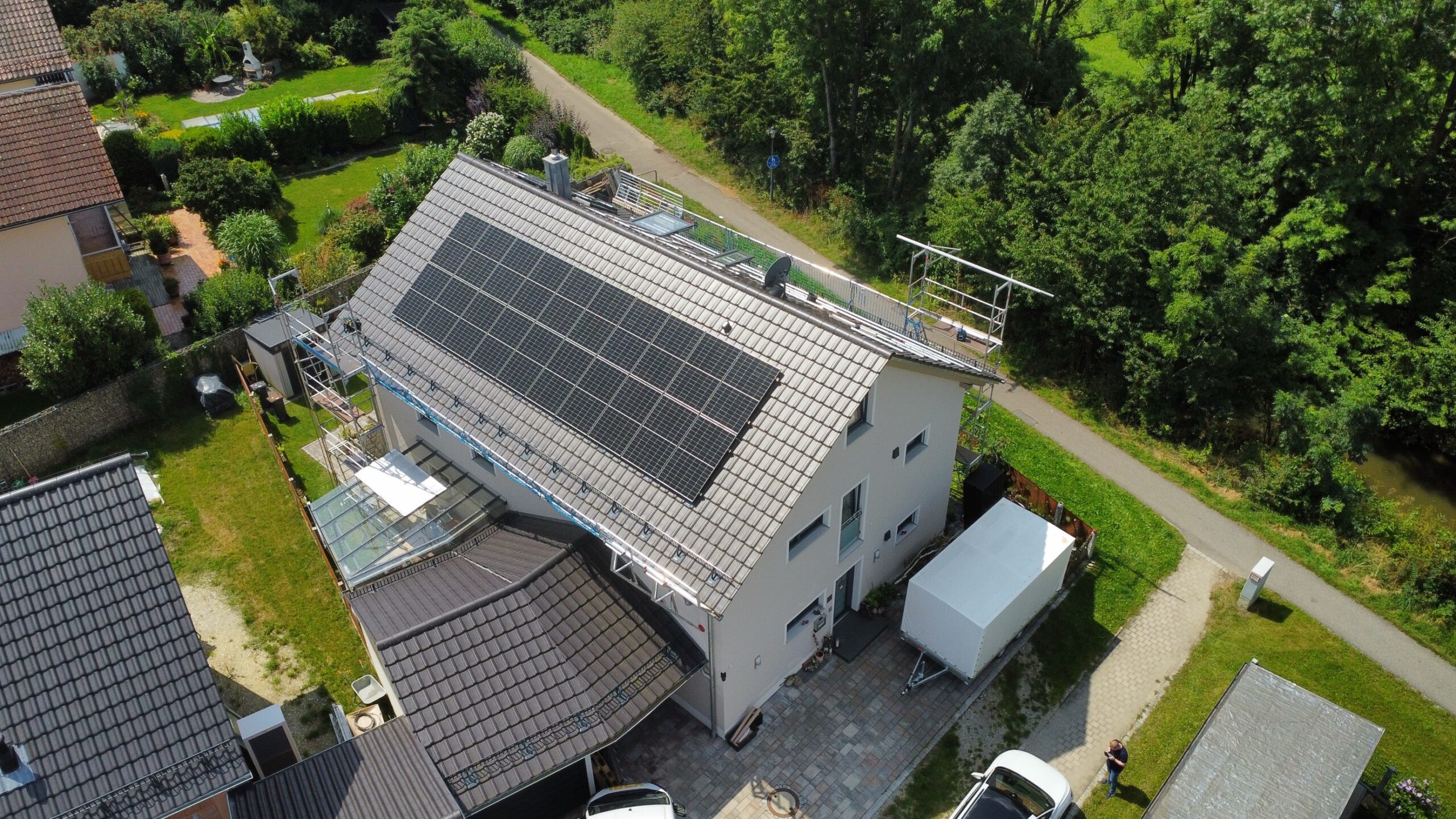 oberbayern-1 Luftaufnahme eines modernen Hauses mit großen Sonnenkollektoren auf dem Dach, umgeben von Grünflächen und Nachbarhäusern, mit einer Einfahrt, geparkten Fahrzeugen und einem Spazierweg in der Nähe.