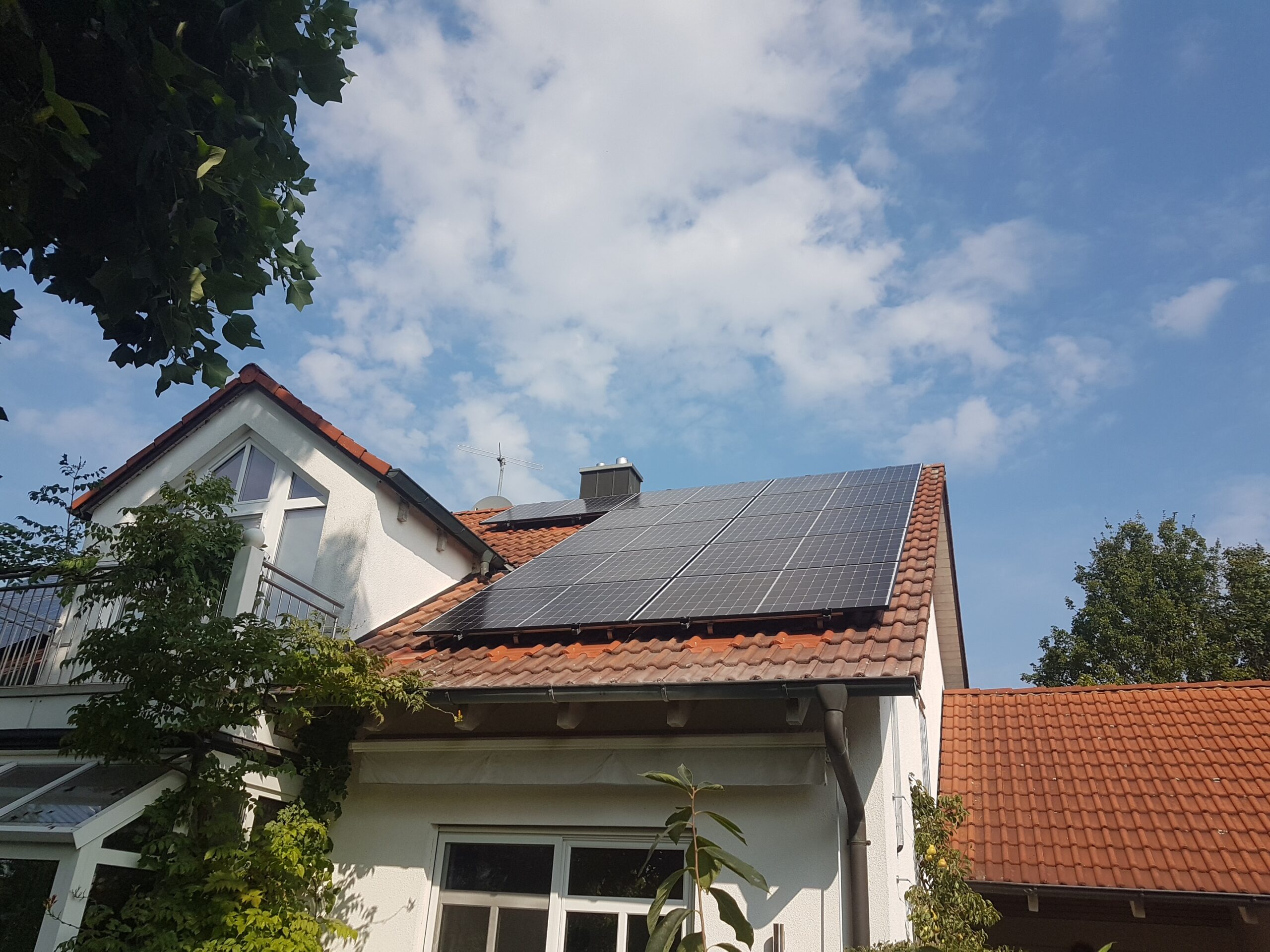 oberbayern-2 Weißes Haus mit rotem Ziegeldach und Sonnenkollektoren, umgeben von Grünflächen. Der Himmel ist blau mit vereinzelten Wolken.