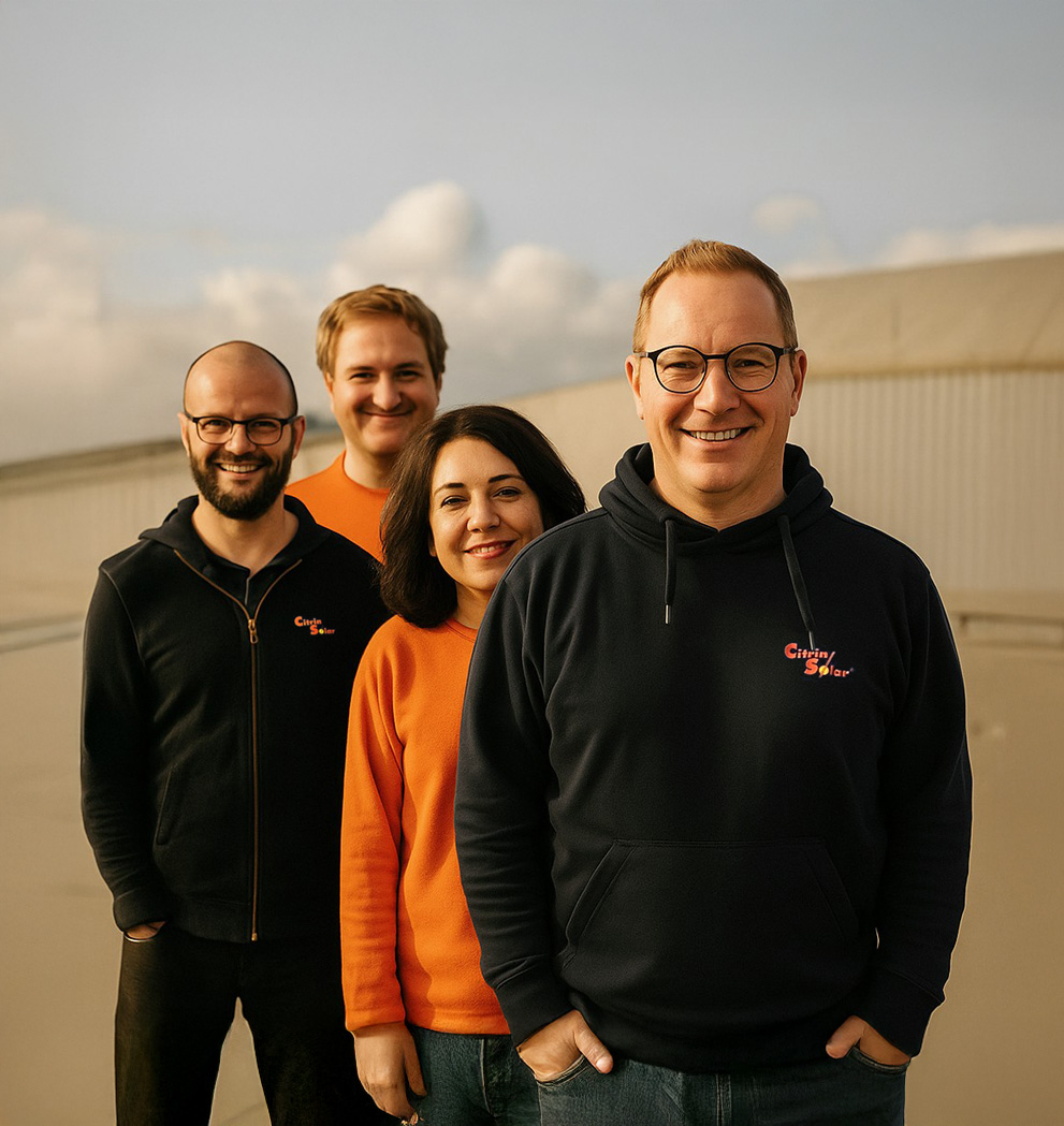 group_image_01 Vier Personen stehen in einer Reihe im Freien und lächeln in die Kamera. Drei Männer und eine Frau tragen legere Kleidung in Orange und Schwarz, im Hintergrund sind ein bewölkter Himmel und ein helles Gebäude zu sehen.